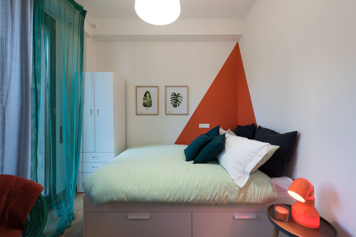 A bedroom with a bed, pillows, a nightstand with a lamp, a small table with a candle, and a wall with two framed plant pictures.
