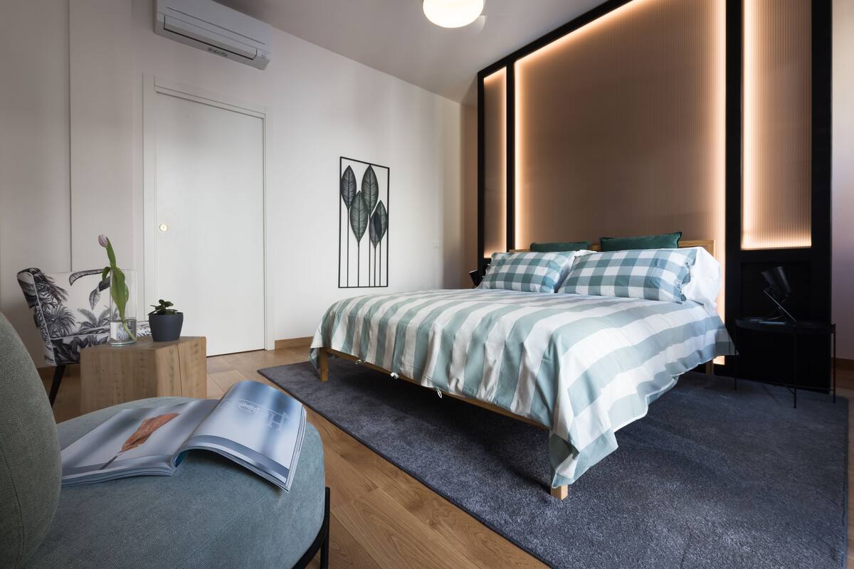 A modern bedroom with a bed, bedside tables, a reading chair, and a wall-mounted air conditioner.