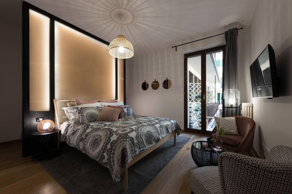 A modern bedroom with a large bed, a hanging light fixture, a TV mounted on the wall, and a sliding glass door leading to a balcony.