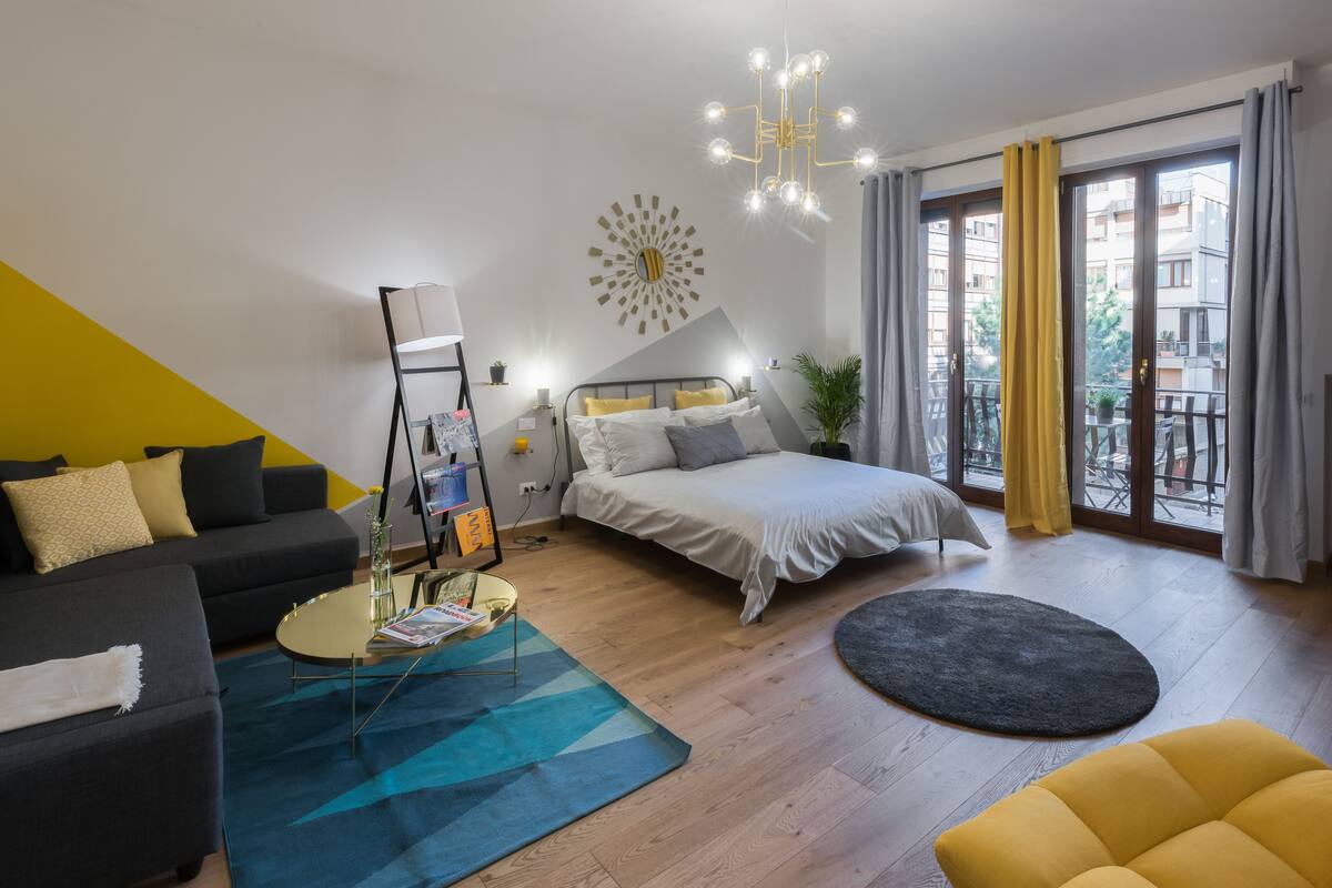 A modern hotel room with a bed, sofa, and a dining area.