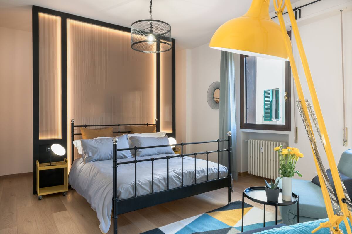 A modern bedroom with a metal bed frame, a bedside table with a lamp, a window with blinds, and a radiator.