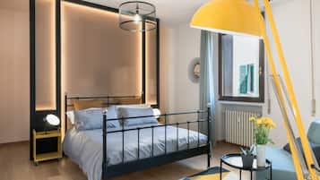Quarto moderno com estrutura de cama em metal, mesa de cabeceira com abajur, janela com persianas e radiador.