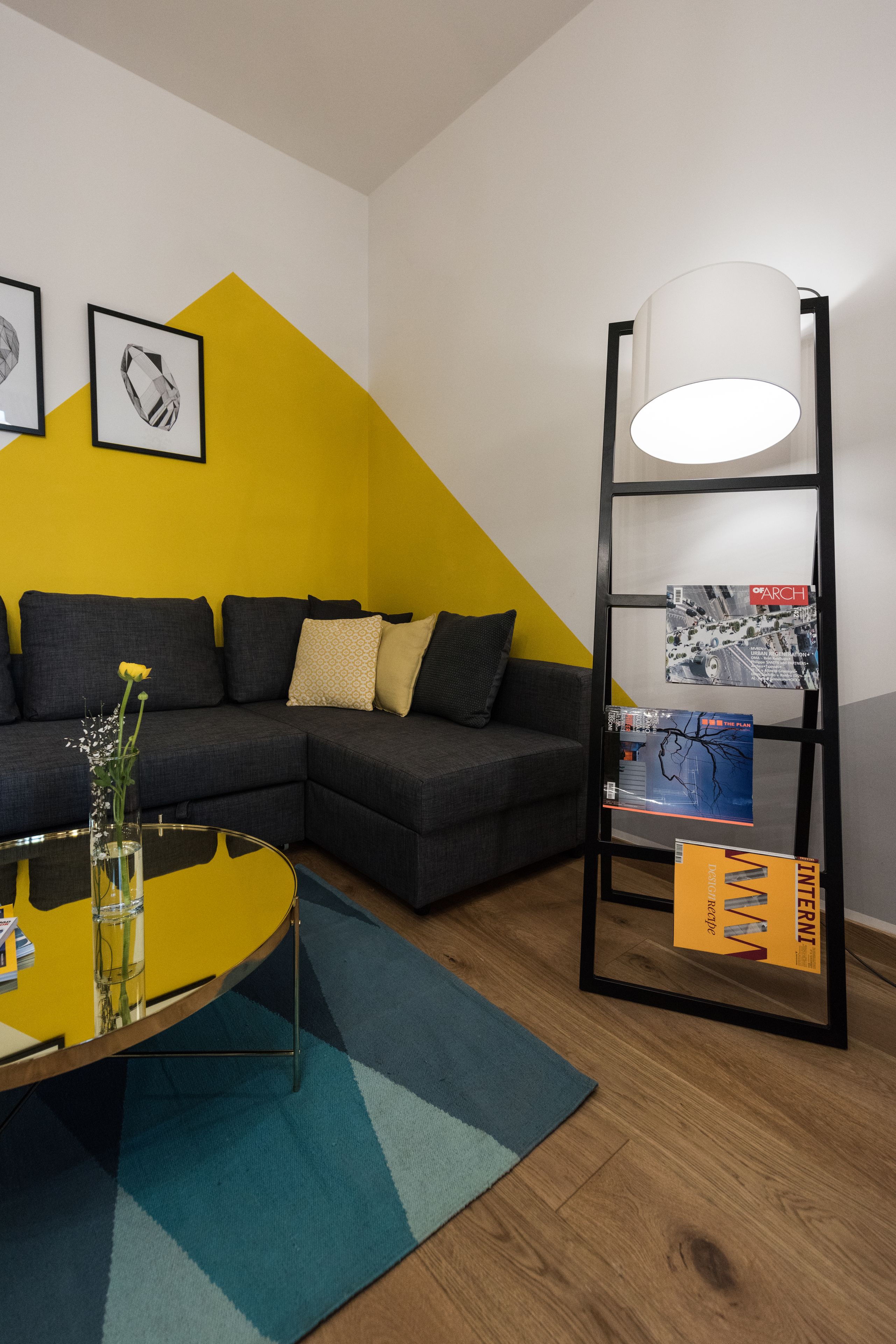 Uma sala de estar moderna com sofá seccional, mesa de centro de vidro e uma estante preta com quadros e uma luminária.