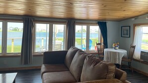 Een kamer met een houten plafond, een bruine leren bank, een eettafel met stoelen en grote ramen met uitzicht op een wateroppervlak.
