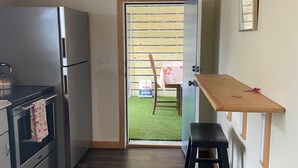 Een compacte keuken met een koelkast, fornuis en een houten plafond. Een klein krukje en een houten plank zijn zichtbaar.