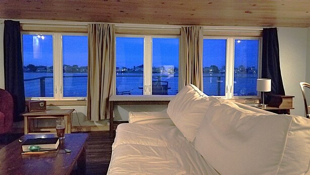 Een hotelkamer met een bed, een bureau, een stoel en uitzicht op een waterpartij door grote ramen.