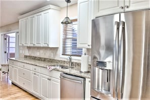 Una cocina moderna con armarios blancos, electrodomésticos de acero inoxidable, encimera de granito y una ventana con persianas.