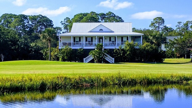 A large, two-story house with a white exterior and a grey roof, surrounded by lush greenery and a pond. - Secluded 3BR private home on golf course! (Edisto Beach)