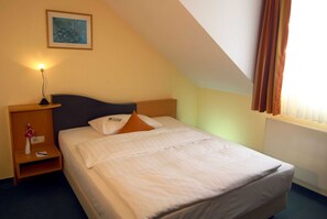 Una habitación de hotel con una cama individual, una mesita de noche con lámpara, un cuadro en la pared y una ventana con cortinas.