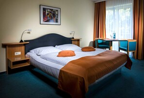 Una habitación de hotel con una cama, dos mesitas de noche, un escritorio con una silla, un cuadro en la pared y una ventana con cortinas.