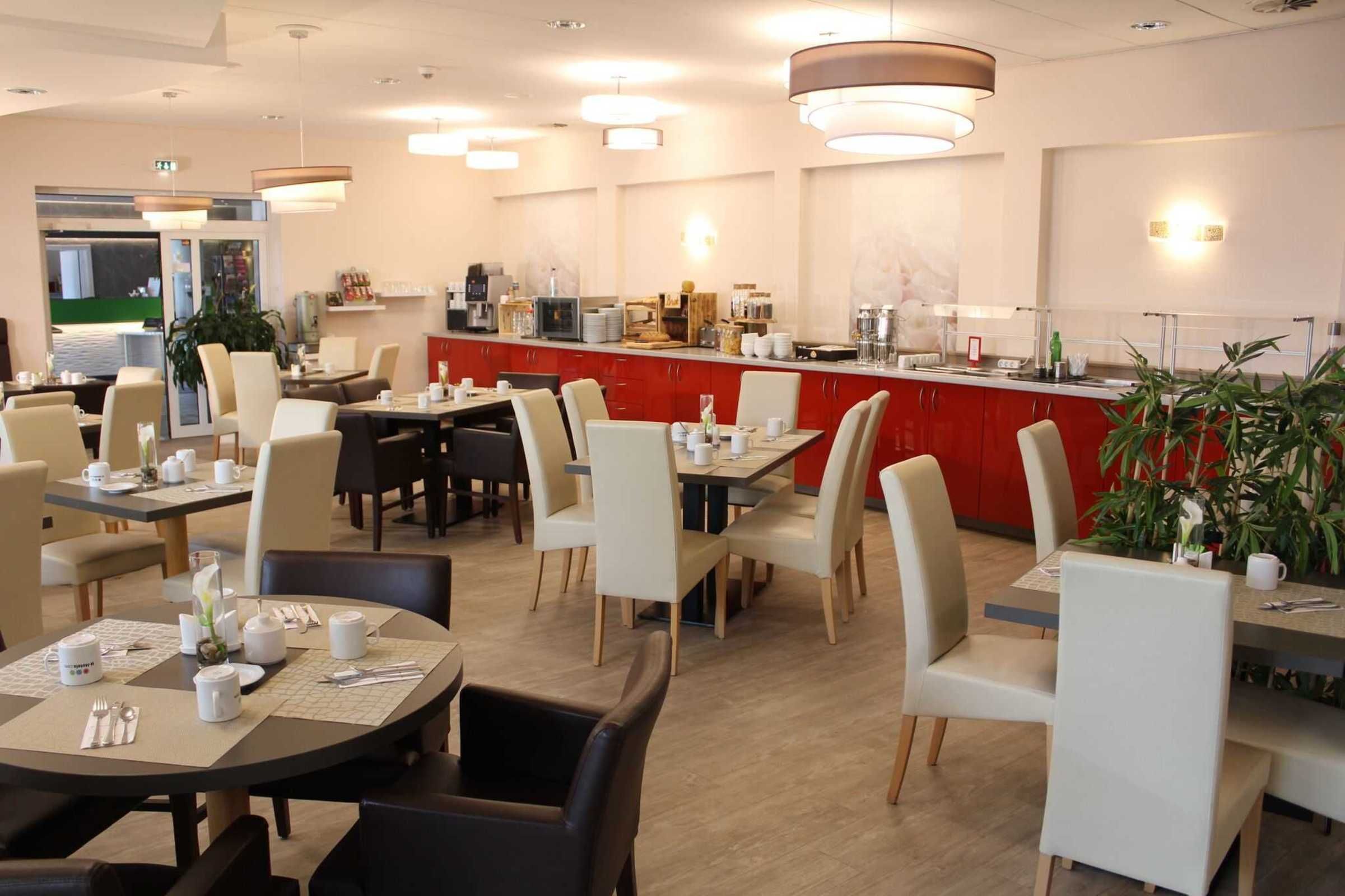 A modern hotel dining area with round tables, beige chairs, and a red counter with coffee machines.