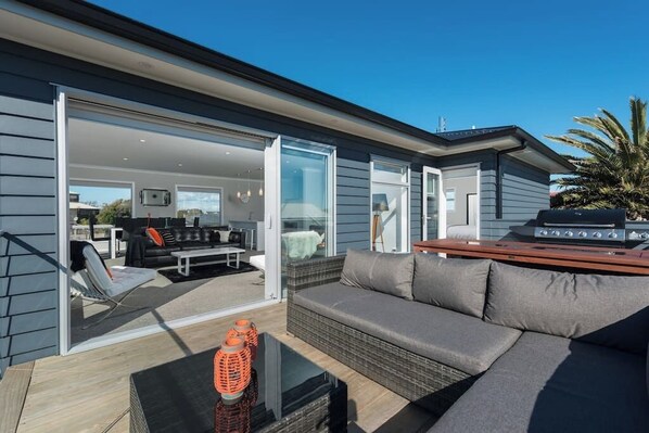 An open-plan living area with a sofa, coffee table, and grill. - Whitesands Beach House - Papamoa Beach (Papamoa)