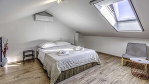Een slaapkamer met een schuine plafond, dakraam, een bed met witte lakens, een nachtkastje, een stoel en een klein rond tafeltje.