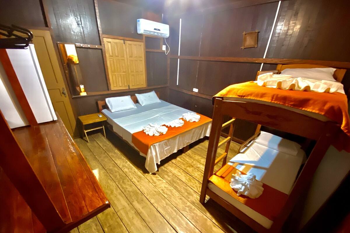 Ein Zimmer mit Holzboden, einem Etagenbett mit orangefarbener Decke, einem Einzelbett mit weißem Bettüberwurf, einem Holztisch, einem kleinen Tisch und einer Wand-Klimaanlage.