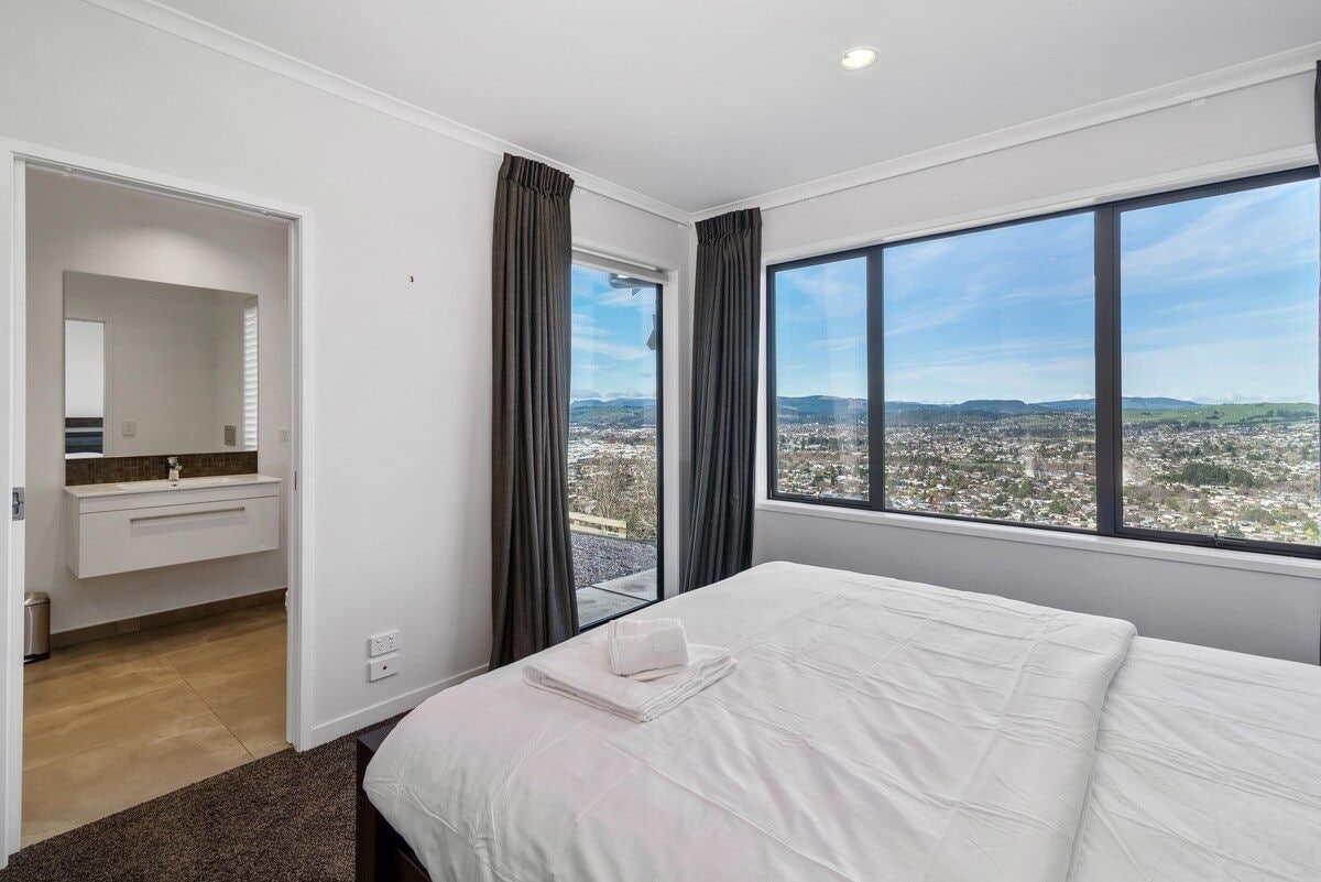 A hotel room with a large bed, a bathroom with a sink and mirror, and a view of a cityscape through large windows.