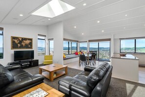 A modern living room with a large window, a flat-screen TV, leather sofas, and a dining area. - The Lookout - Rotorua Views, Spa Pool, Sleeps 7 (Rotorua Township)