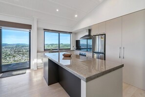 A modern kitchen with a large island, stainless steel appliances, and a view of a cityscape through large windows. - The Lookout - Rotorua Views, Spa Pool, Sleeps 7 (Rotorua Township)