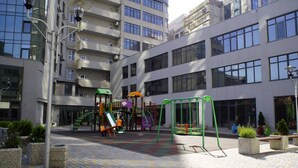En legeplads med forskellige legekonstruktioner, omgivet af en bygning med flere etager og store vinduer.