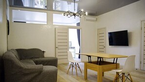 En moderne stue med sofa, spisebord med stole, fjernsyn monteret på væggen og store vinduer der lukker naturligt lys ind.