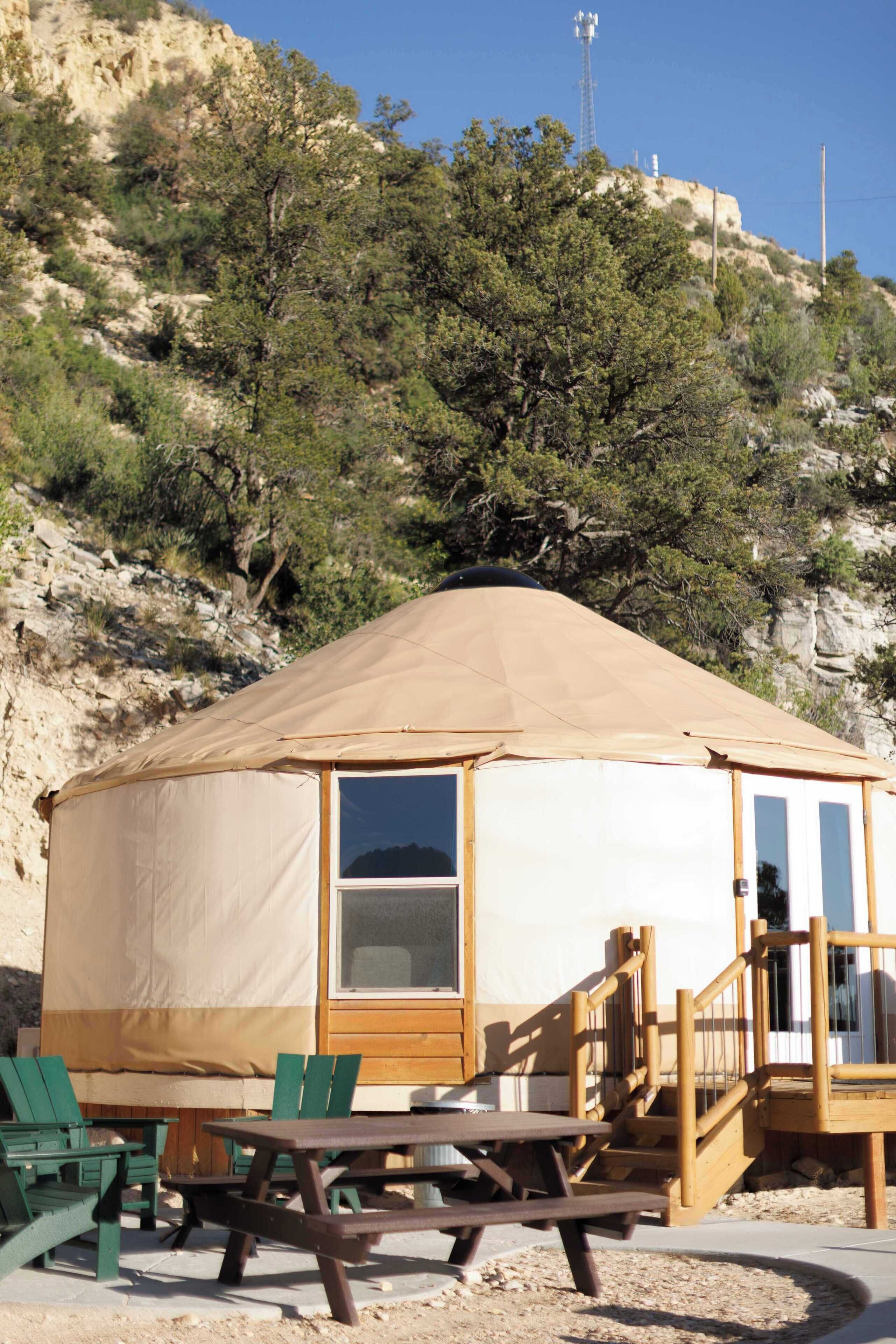 A yurt with a wooden deck, picnic table, and chairs, set against a hillside with trees and a clear sky.