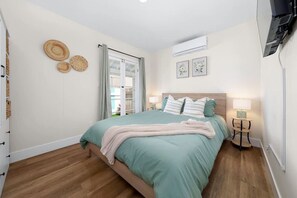 A bedroom with a bed, a bedside table, a lamp, a window with curtains, and wall decorations. - Suite Cozy Beach Nook, 150 Steps To The Sand (San Diego)