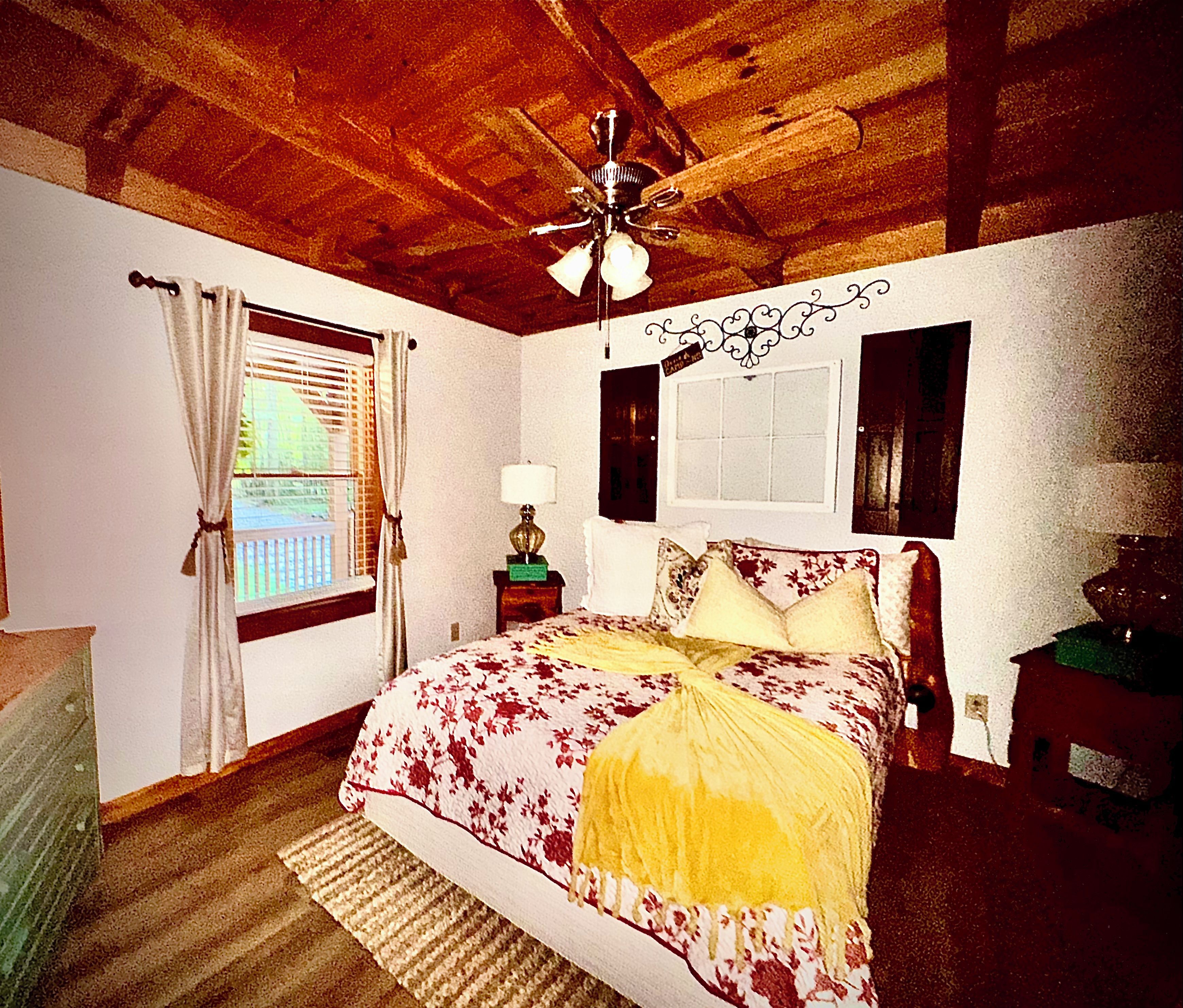 Une chambre avec un plafond en bois, un lit recouvert d’une literie à motifs floraux, une table de chevet avec une lampe et une fenêtre avec des rideaux.