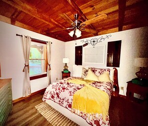 A bedroom with a wooden ceiling, a bed with floral bedding, a nightstand with a lamp, and a window with curtains. - Majestic, private cabin with hot tub/multiple deck...a little taste of heaven! (Dahlonega)