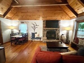 A cozy living room with a fireplace, wooden ceiling, and a dining area with a table and chairs.