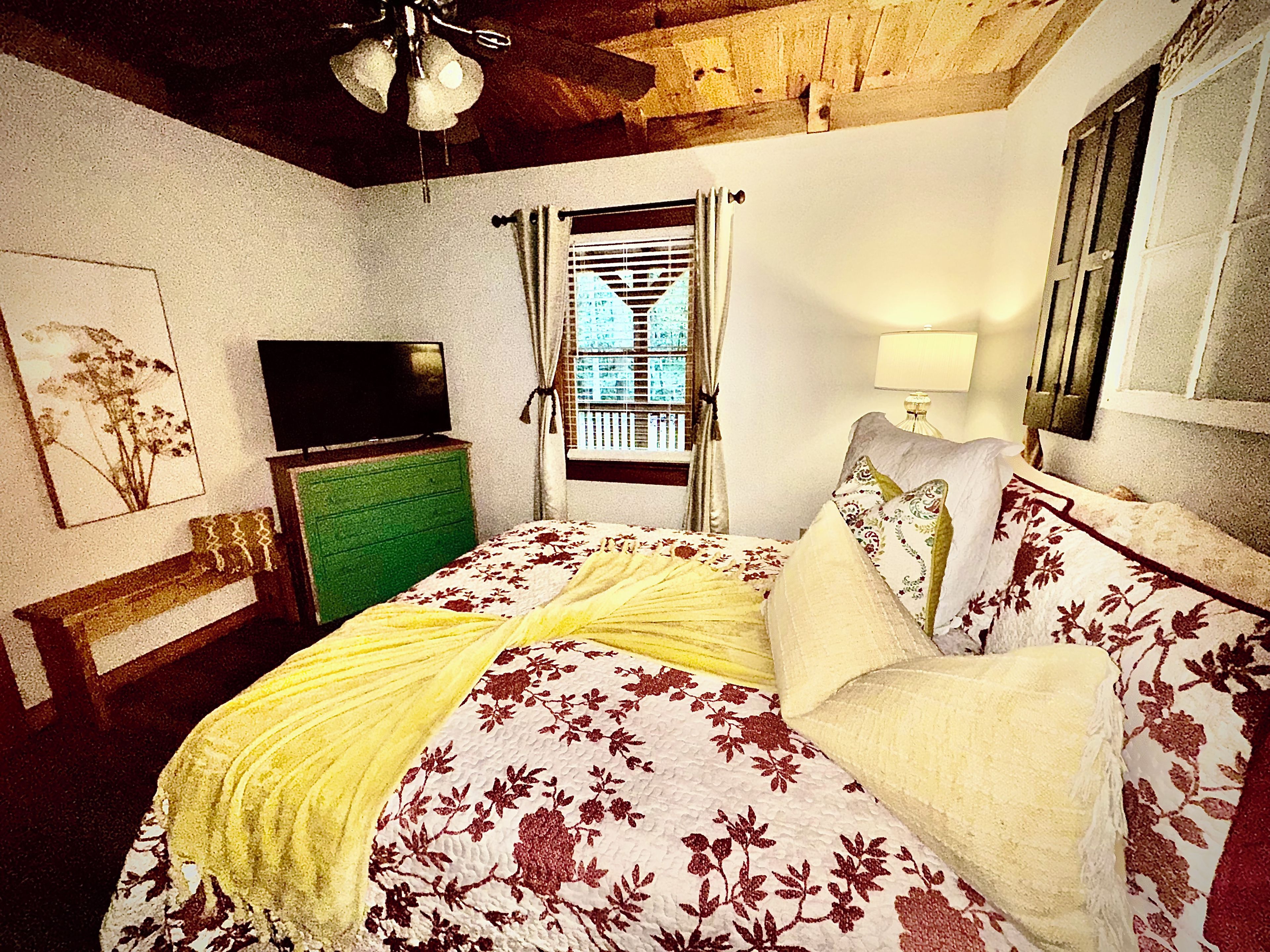 A bedroom with a bed, a television on a dresser, a window with curtains, and a lamp.