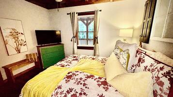A bedroom with a bed, a television on a dresser, a window with curtains, and a lamp.