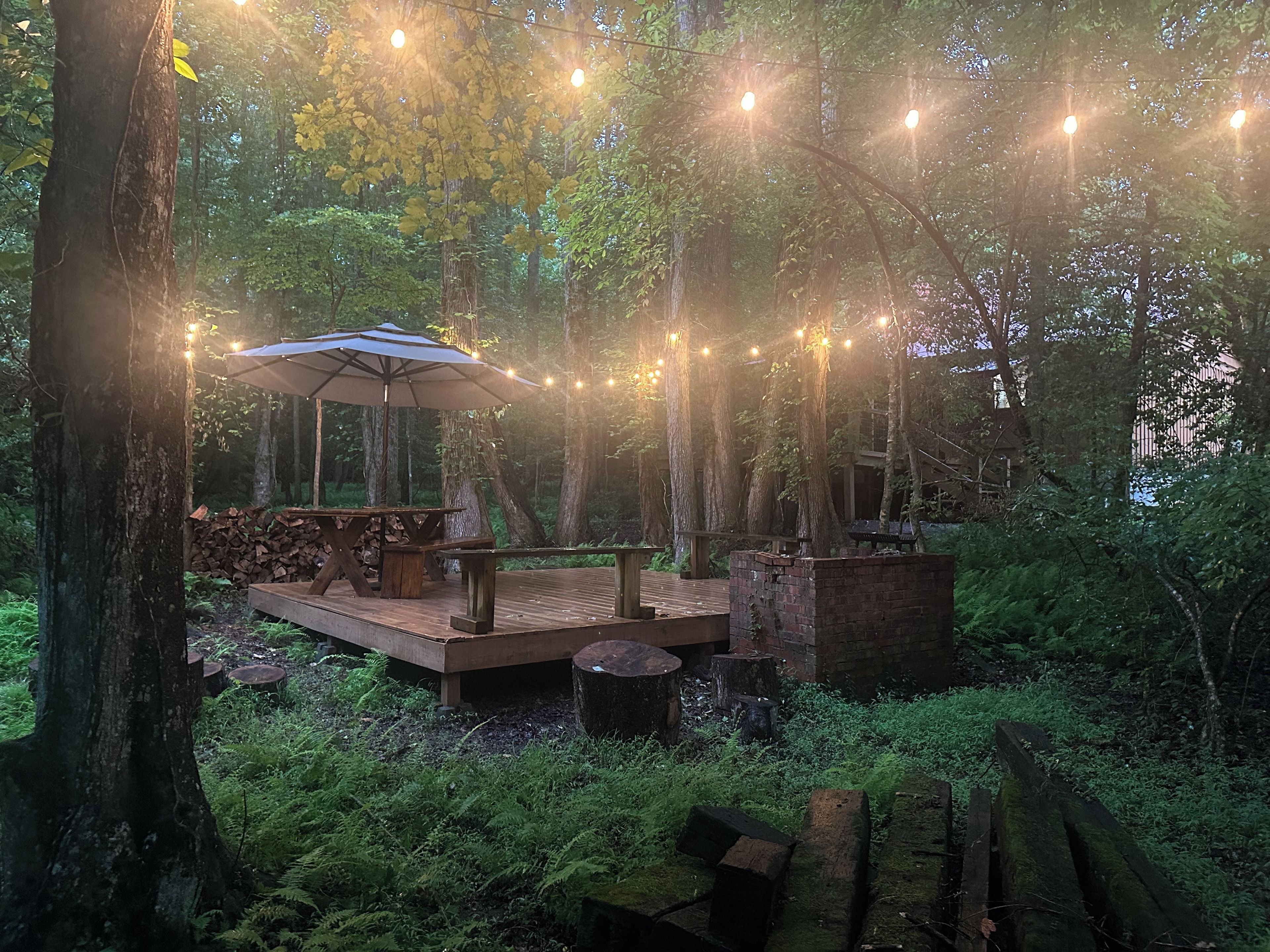 A wooden deck with a table and bench, surrounded by trees and string lights.
