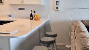 A kitchen with a white countertop, a 'NO SMOKING' sign, a wine bottle, and a basket of bananas. There are three stools and a couch in the background.
