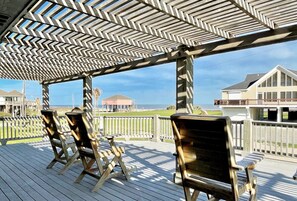 A wooden deck with a chevron-patterned pergola, overlooking a beach and a house.