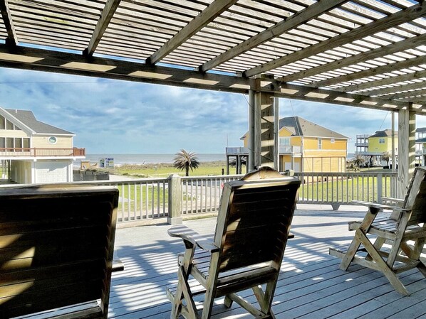 A covered outdoor area with wooden chairs and tables, overlooking a beach and ocean.