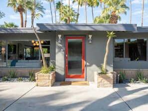Une maison moderne avec une porte rouge, des piliers en pierre et une vue sur l’intérieur grâce à de grandes fenêtres.