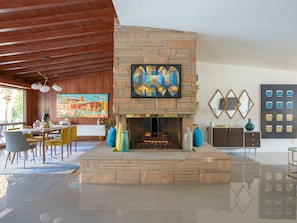 A spacious living room with a stone fireplace, wooden ceiling, and a dining area with yellow chairs.