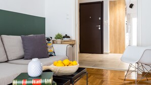 A modern living room with a sofa, a coffee table with a bowl of oranges, a white vase, and a book. There is a wooden door and a white chair in the background. - Special Prices Urban Athens Apartment 2 min walk from metro (Athina)