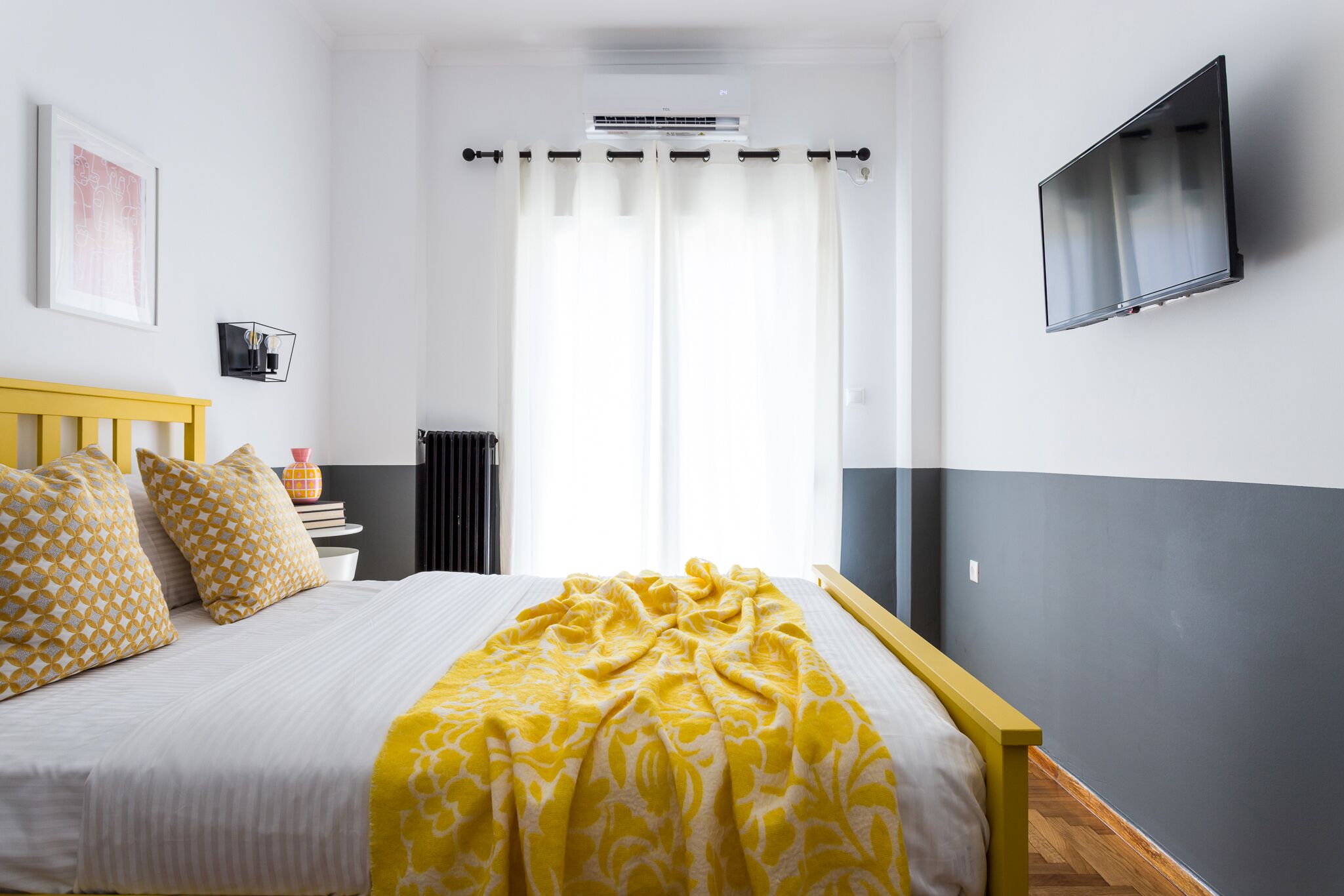 A bedroom with a bed, a bedside table, a wall-mounted TV, and a window with curtains.