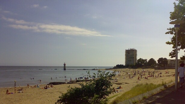 A beach with people, a lighthouse, and a tall building in the background. - New apartment for rent with luxurious furnishings (Saint-Nazaire)