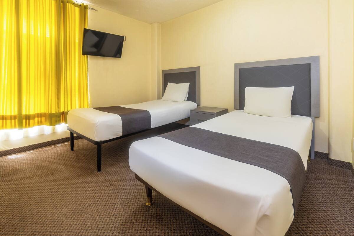 A hotel room with two single beds, a flat-screen TV mounted on the wall, and a window with yellow curtains.
