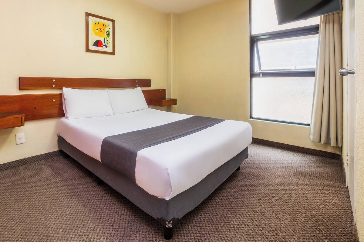 A hotel room with a bed, a wooden headboard, a small bedside table, a wall-mounted TV, a window with curtains, and a framed picture on the wall.
