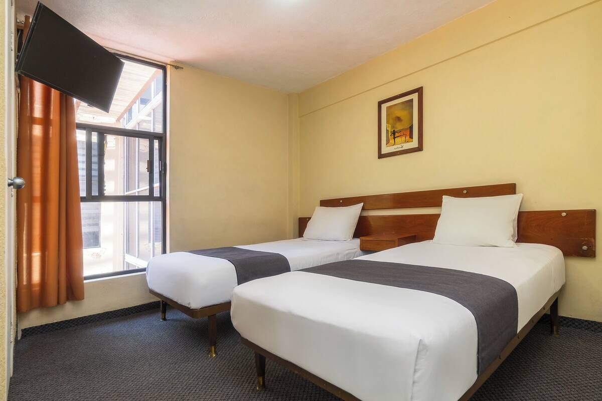 A hotel room with two beds, a large window with curtains, a flat-screen TV mounted on the wall, and a framed picture above the beds.