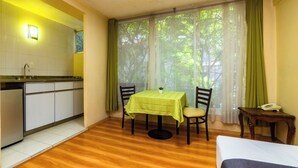 A compact hotel room with a wooden floor, a small dining table with a green tablecloth, two chairs, a single bed, a bedside table with a phone, and a window with sheer curtains. - Capital O Hotel Suites Marne, Mexico City (Mexico City)