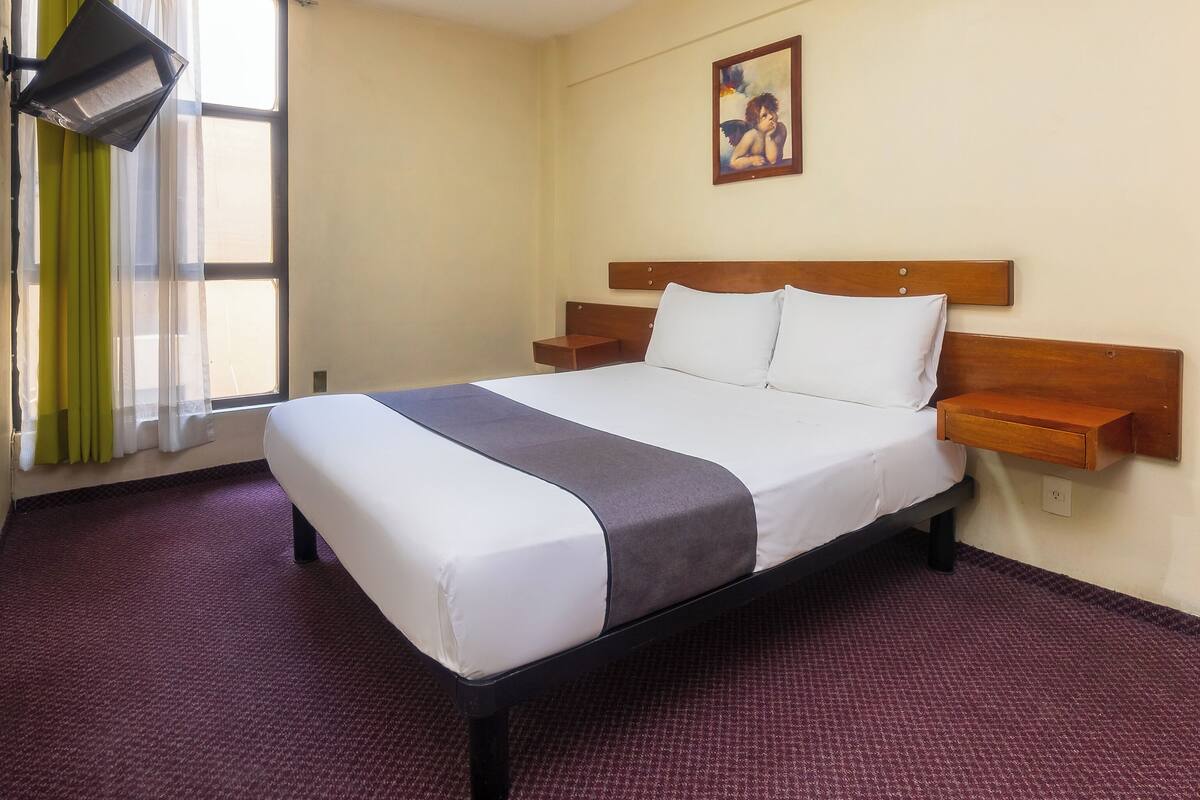 A hotel room with a bed, a nightstand, a wall-mounted lamp, and a painting.