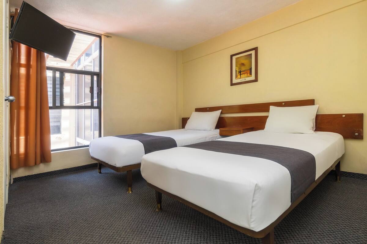 A hotel room with two beds, a television, a window with curtains, and a framed picture on the wall.