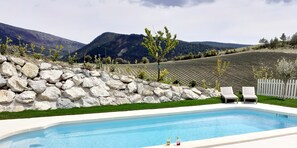A swimming pool with two lounge chairs, a stone wall, and a view of a vineyard and mountains. -  Holiday rental Gite Pousada Tropical (VOLVENT)