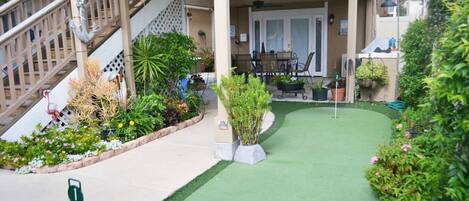 A residential property with a covered patio, a staircase, and a small garden area with various plants and a green artificial turf.