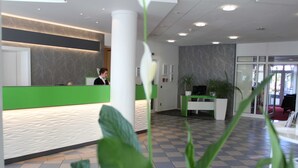 Aula, jossa on vihreä vastaanottotiski, valkoinen pylväs ja ruutukuviollinen lattia.