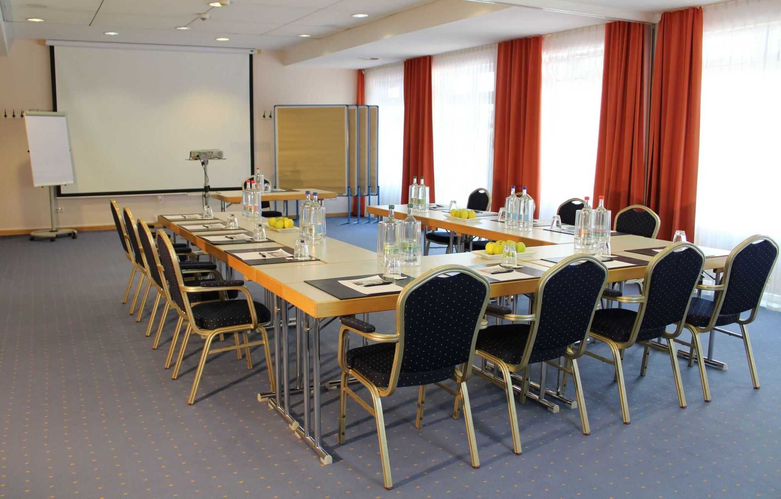 Ein Konferenzraum mit einem langen Tisch, Stühlen, einer Projektionsleinwand und einer Whiteboard-Tafel.