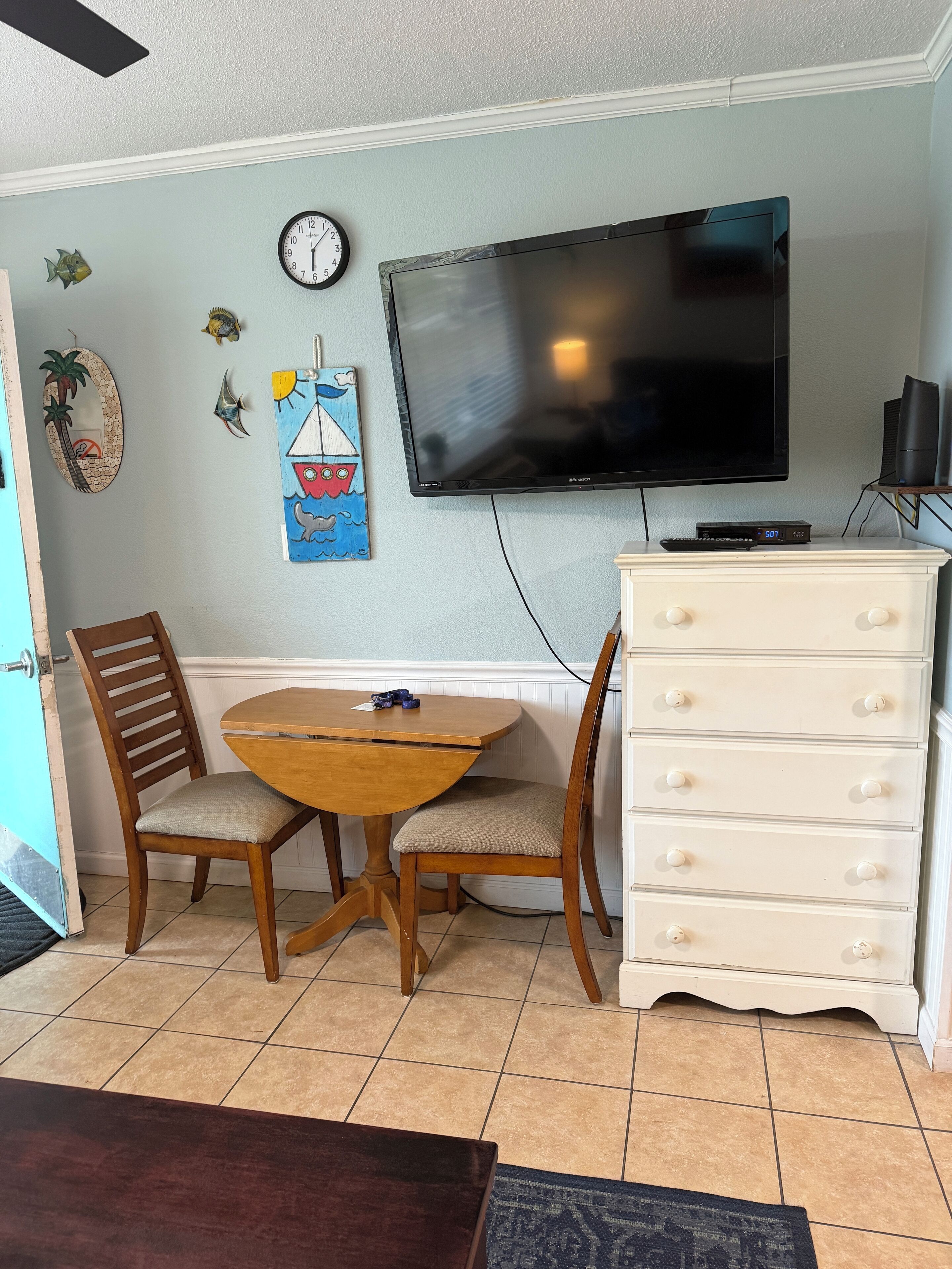 Un coin repas avec une table et des chaises en bois, une commode blanche, un téléviseur à écran plat fixé au mur et une horloge.